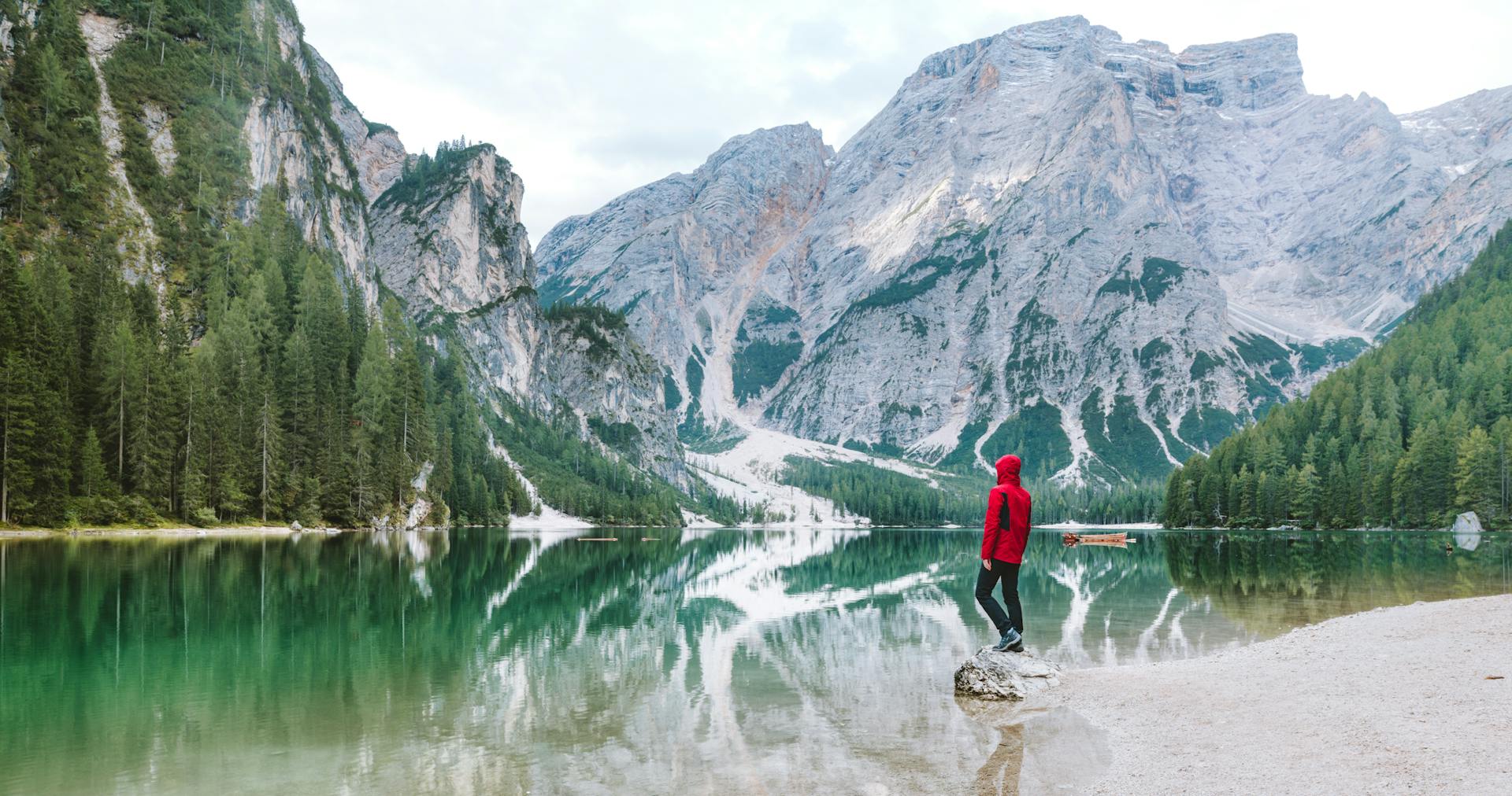 A person in a red jacket stands on a rock by a tranquil lake, surrounded by towering mountains and lush green trees, reflecting in the water.